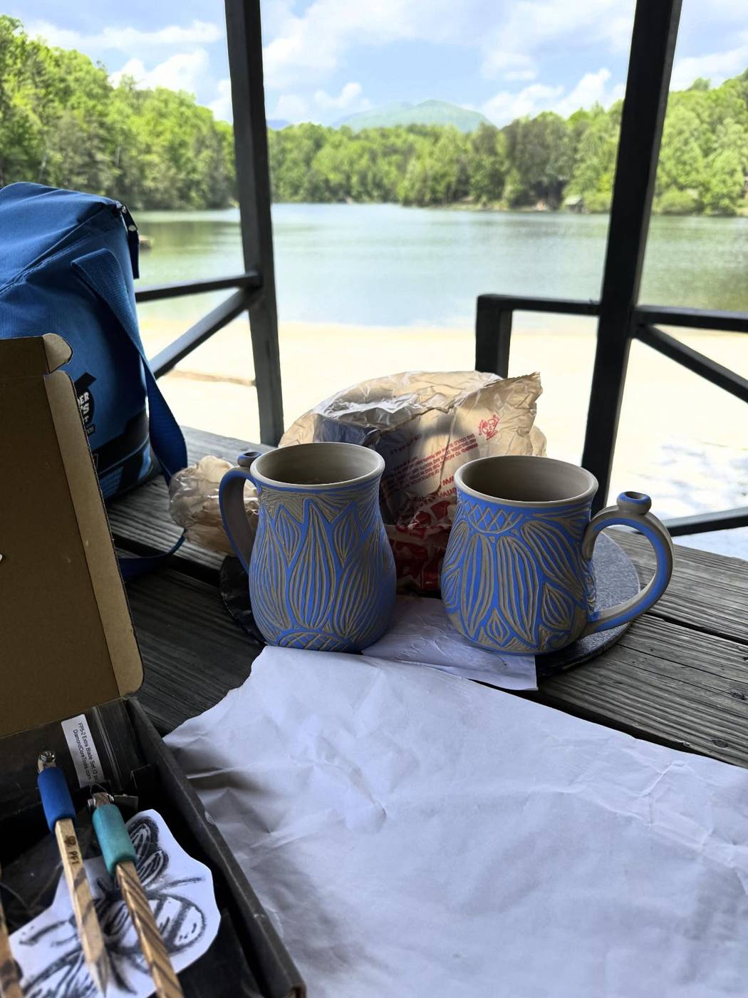 Two blue carved mugs on a pavilion railing, a lake and wooded hills behind them